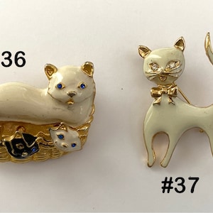 May include: Two white enamel cat brooches with blue rhinestone eyes. The first brooch features a cat with three kittens in a basket. The second brooch features a cat with a bow tie. The brooches are numbered #36 and #37.