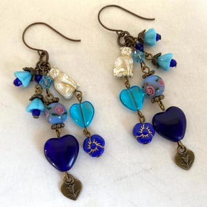 May include: A pair of dangle earrings with a whimsical design. Each earring features a dark bronze hook, with blue bell-shaped beads, white cat charms, and heart-shaped beads in turquoise and deep blue. The earrings are adorned with floral and leaf charms.