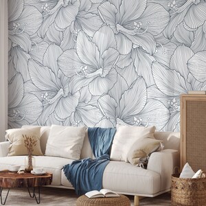 Peut inclure: Un canap&eacute; blanc avec des plaids bleus devant un mur avec un motif floral blanc et gris. Le canap&eacute; a deux coussins et une table basse en bois avec un vase de fleurs dessus.