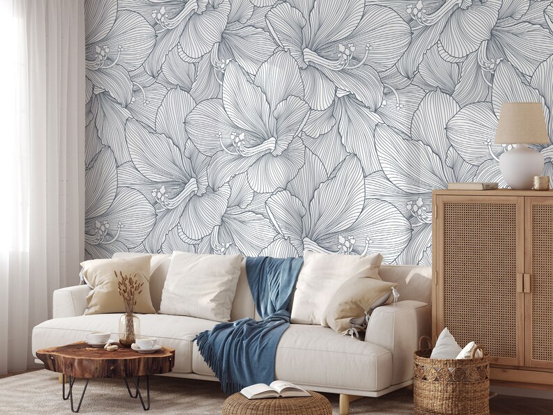 Peut inclure: Un canap&eacute; blanc avec des plaids bleus devant un mur avec un motif floral blanc et gris. Le canap&eacute; a deux coussins et une table basse en bois avec un vase de fleurs dessus.