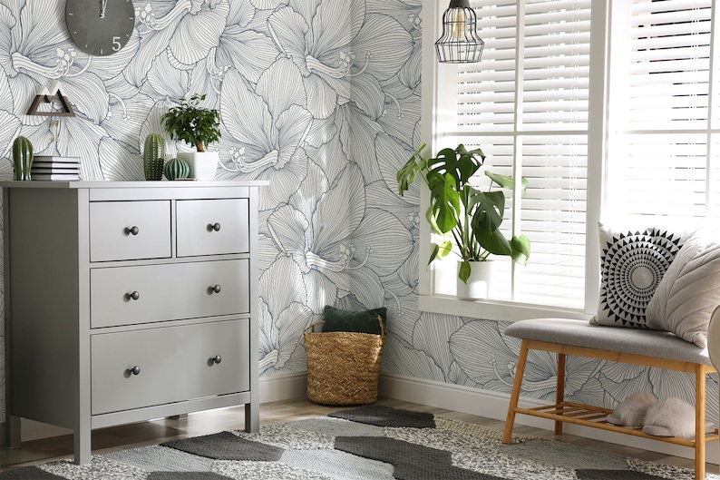 Peut inclure: Une commode grise &agrave; quatre tiroirs et des boutons noirs se trouve dans une pi&egrave;ce avec un mur blanc orn&eacute; d'un papier peint &agrave; motifs floraux. Une plante en pot est pos&eacute;e sur la commode. Un panier en osier se trouve sur le sol &agrave; c&ocirc;t&eacute; d'un banc gris avec un coussin et un oreiller &agrave; motifs noir et blanc.