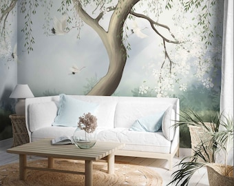 May include: A white couch with blue pillows sits in front of a wall mural depicting a large tree with white blossoms and birds flying in the sky. A wooden coffee table with a book and a glass vase with flowers sits in front of the couch. A woven rug is on the floor.