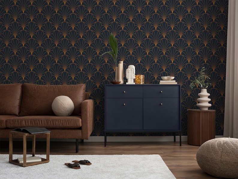 K&ouml;nnte beinhalten: Eine dunkelblaue Tapete mit einem goldenen geometrischen Muster aus &uuml;berlappenden F&auml;cherformen. Vor der Tapete steht ein braunes Ledersofa mit einem Holz-Beistelltisch und einem Buch darauf. Rechts vom Sofa steht ein dunkelblauer Schrank mit zwei Schubladen und zwei T&uuml;ren. Rechts vom Schrank steht ein Holz-Beistelltisch mit einer beigefarbenen Vase und einem beigefarbenen Ottoman.