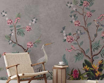 May include: A gray wall with a floral wallpaper featuring pink flowers, green leaves, and birds. A white crane is depicted on the left side of the wall. A wicker chair and a side table with a book and a cup are in the foreground.