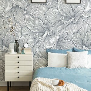 Peut inclure: Une chambre &agrave; coucher avec un papier peint floral blanc et bleu, une table de chevet blanche &agrave; quatre tiroirs, un lit avec une couette bleue et une couverture en tricot blanc, et deux dessins encadr&eacute;s sur le mur.