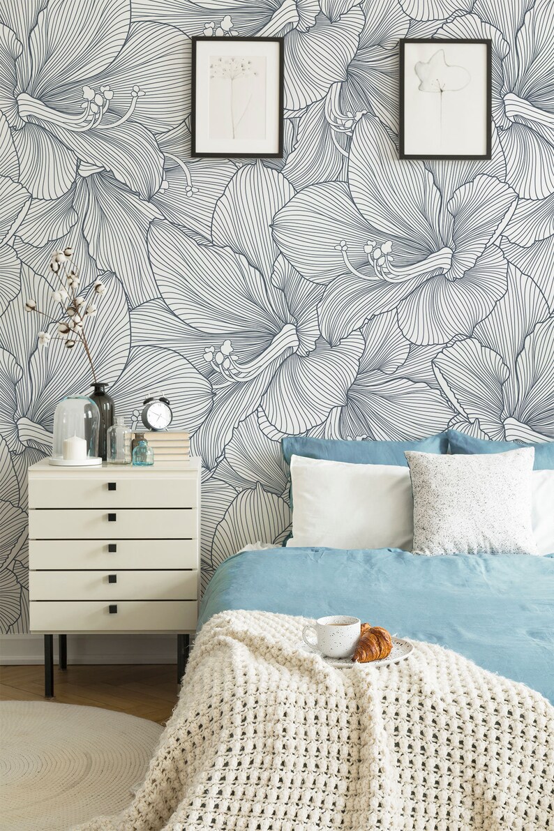 Peut inclure: Une chambre &agrave; coucher avec un papier peint floral blanc et bleu, une table de chevet blanche &agrave; quatre tiroirs, un lit avec une couette bleue et une couverture en tricot blanc, et deux dessins encadr&eacute;s sur le mur.