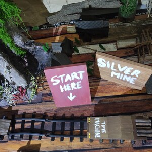 May include: A miniature train set with a wooden 'Time Machine' carriage on a track. The set is on a wooden surface with a red sign that says 'Start Here' and a brown sign that says 'Silver Mine'.