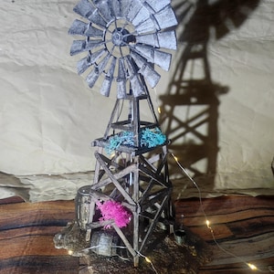 May include: A miniature wooden windmill with a weathered gray paint finish and a teal and pink moss decoration. The windmill is on a wooden base with a string of fairy lights.