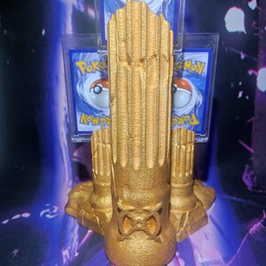 Könnte beinhalten: Ein goldfarbener, 3D-gedruckter Ständer mit einem strukturierten, säulenartigen Design. Der Ständer ist so konzipiert, dass er Sammelkarten aufnehmen kann, mit Steckplätzen für mehrere Karten. Der Ständer hat eine Basis mit einem dekorativen Design.