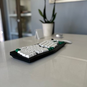 Könnte beinhalten: Eine schwarz-weiße mechanische Tastatur mit grünen Akzenten. Die Tastatur hat weiße Tasten und einen schwarzen Rahmen. Die Tastatur liegt auf einer weißen Oberfläche. Eine weiße Maus befindet sich neben der Tastatur.