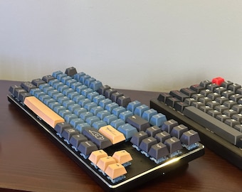 May include: Two mechanical keyboards with keycaps in shades of blue, tan, and black. One keyboard has a red escape key. The keyboards are on a dark wooden surface. The keycaps have a variety of symbols and letters.