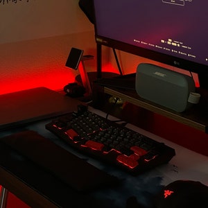 May include: A black mechanical keyboard with red backlit keys sits on a desk, next to a mouse and a mousepad with a blue and white design. A monitor displays text, and a Bose speaker is visible.
