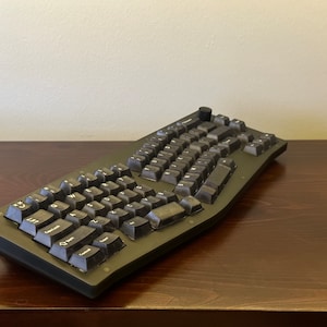 May include: A black ergonomic keyboard with a curved design and dark keycaps. The keyboard has a unique layout with split sections and a central control knob. It sits on a dark wooden surface.