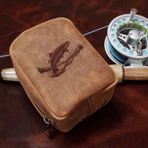 May include: A brown leather fishing reel case with a fish graphic, a fishing rod, and a silver fishing reel with blue and green line. The case is rectangular with a zipper closure.