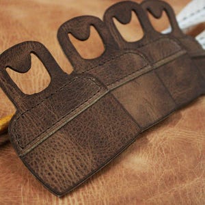 May include: A brown leather archery finger tab with four finger loops. The tab is textured and stitched, with a worn appearance. Two arrows with white fletching are visible. A wooden arrow is also present.