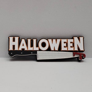 May include: A Halloween-themed decorative item featuring the word "HALLOWEEN" in white letters with an orange outline, above a knife with a silver blade and a red blood-like stain on the tip.