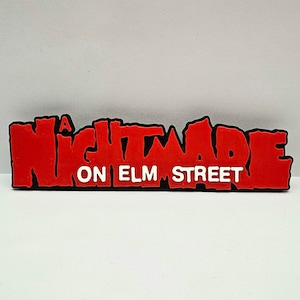 Op de afbeelding: Een rood en zwart logo met de tekst "A Nightmare on Elm Street" in witte letters.