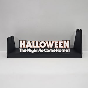 May include: A black Halloween-themed shelf with the word "HALLOWEEN" in white block letters. Below the word is the phrase "The Night He Came Home!" in white letters. The shelf has two upright supports.