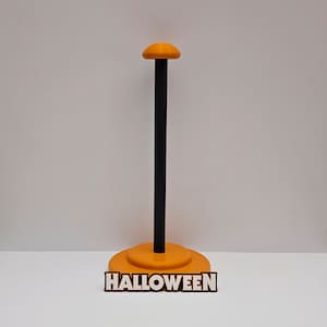 May include: Orange Halloween head stand with a black pole and a round base. The base has the word "HALLOWEEN" printed on it.