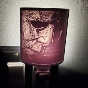 Puede incluir: Una luz nocturna morada con una imagen blanca y gris de una cara de miedo. La luz nocturna se conecta a una toma de corriente estándar.