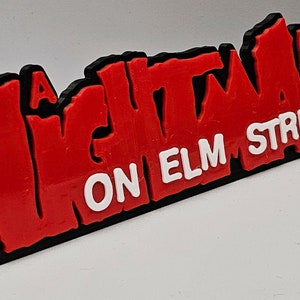 Op de afbeelding: Een "A Nightmare on Elm Street" logo, rood met zwarte omtrek, op een witte achtergrond. De tekst is in een vet, gestileerd lettertype.