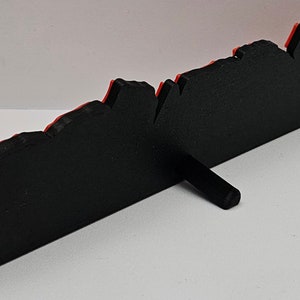 Op de afbeelding: Zwarte en rode 3D-geprinte wanddecoratie met een grillig, bergachtig ontwerp. De decoratie heeft een klein standaard aan de achterkant voor weergave.