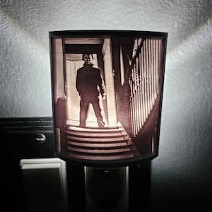 Puede incluir: Una luz nocturna con una imagen impresa en 3D de un hombre en un pasillo oscuro. La imagen está en tonos grises y marrones.