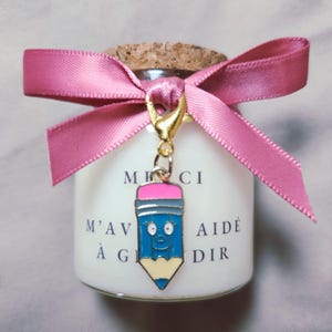 Peut inclure: Un petit pot blanc avec un couvercle en liège, noué avec un ruban rose. Un charm doré en forme de crayon bleu avec un visage souriant est suspendu au ruban. Le pot porte du texte.