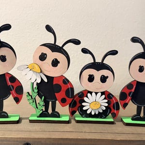 May include: Four wooden ladybug figurines with black bodies, red spotted wings, and smiling faces. Each ladybug holds a daisy or is adorned with a daisy. The figurines stand on green bases, creating a charming spring or garden theme.