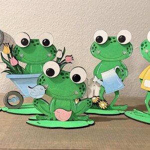 May include: A collection of six green frog cutouts with large eyes, each on a green lily pad base. One frog holds a butterfly net, another a watering can, and a third a pink and blue umbrella. Other frogs have a butterfly, a bird, and a wheelbarrow.