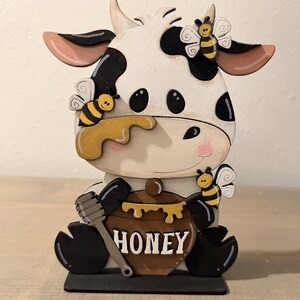 May include: A wooden cow figurine with black and white spots, large black eyes, and pink cheeks. The cow holds a brown honey pot with the word "HONEY" and a honey dipper. Yellow and black bees are attached to the cow's head and face.