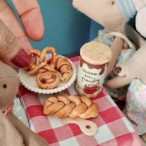 May include: A miniature set featuring a white scalloped plate with a pretzel, croissant, and cinnamon roll, a cylindrical container labeled cacao en poudre with a red mug graphic, and a braided bread loaf on a small wooden cutting board.