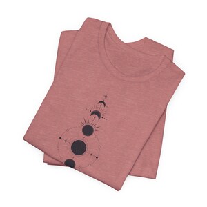 May include: A folded heather pink t-shirt with a black and white graphic of a celestial design featuring moons and stars.