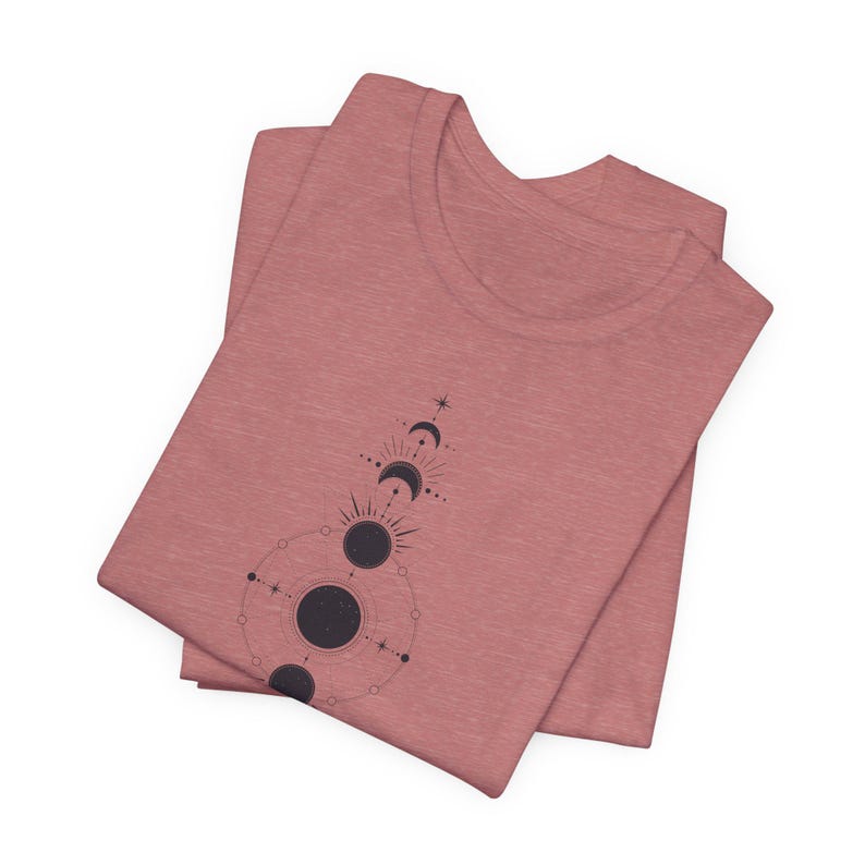May include: A folded heather pink t-shirt with a black and white graphic of a celestial design featuring moons and stars.
