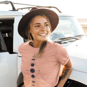 May include: A woman wearing a blush pink t-shirt with a black and white moon phase graphic design. She is wearing a brown hat and looking to the right.