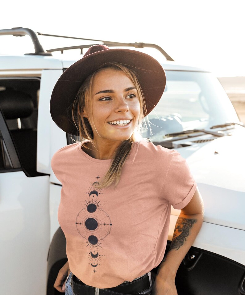 May include: A woman wearing a blush pink t-shirt with a black and white moon phase graphic design. She is wearing a brown hat and looking to the right.