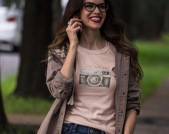 May include: A woman wearing a pink t-shirt with a vintage camera graphic and the text "Snap That" printed on it. She is wearing a brown jacket over the shirt and has long brown hair.