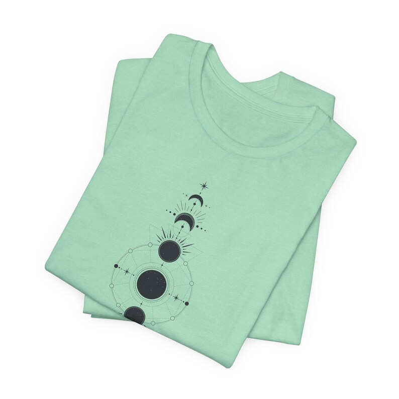May include: A light green t-shirt featuring a black and white geometric design of a crescent moon and sun.
