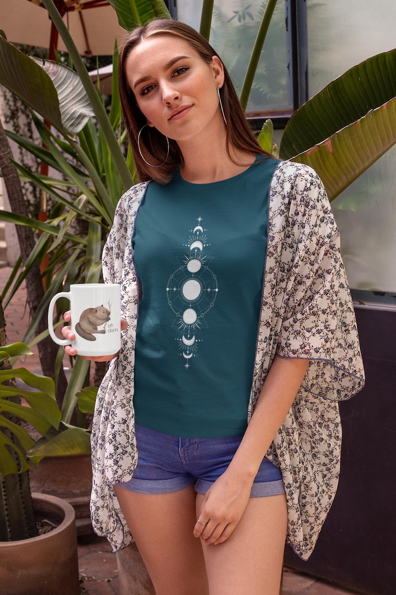 May include: A woman wearing a teal blue t-shirt with a white moon phases design. She is holding a white mug with a brown beaver graphic and the text "Oh, darn".
