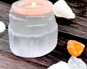 May include: A white selenite candle holder with a lit tea light inside. The holder is made of natural selenite crystals and has a rough, textured surface. There are several small selenite crystals and orange stones scattered around the candle holder on a wooden surface.