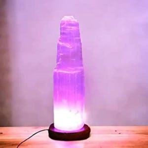 Könnte beinhalten: Eine hohe, weiße Selenitkristall-Lampe mit einem Holzsockel. Die Lampe ist mit einem violetten Licht beleuchtet.
