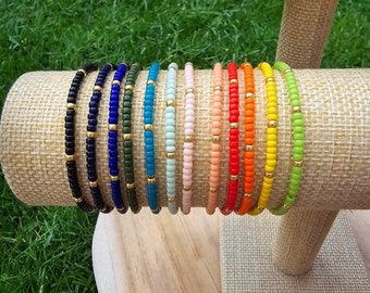 May include: A display of 15 colorful beaded bracelets with gold accents. The bracelets are arranged in a rainbow pattern on a burlap-covered stand.