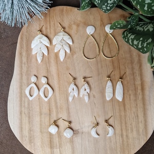 May include: A set of 10 white and gold earrings with various shapes, including leaves, teardrops, and geometric designs. The earrings are displayed on a wooden surface.