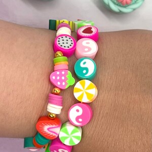 Puede incluir: Un brazalete colorido con varios encantos, incluyendo una sandía, un corazón, un símbolo yin yang, una fresa, un diseño de remolino y una cuenta dorada. El brazalete está en la muñeca de una persona.