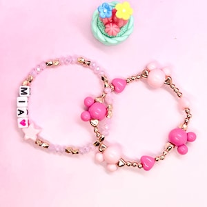 May include: Two beaded bracelets, one with pink and gold beads and the other with clear and gold beads. The clear beaded bracelet has the name "MIA" spelled out with white letter beads.
