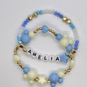 Pode incluir: Um bracelete de contas azuis e brancas com detalhes dourados. O bracelete tem o nome "AMELIA" escrito em contas brancas, juntamente com pingentes de estrelas e cabeças de Mickey Mouse.