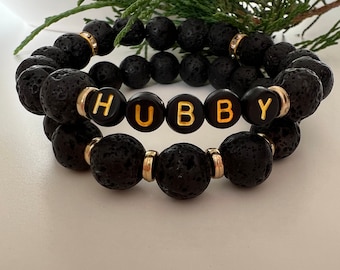 Op de afbeelding: Twee zwarte lava armbanden met gouden kralen en gouden letters die "HUBBY" spellen.