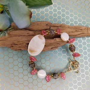 May include: A bracelet with pink and gray beads, a white oval shell, and gold accents. The bracelet has a lobster clasp closure.