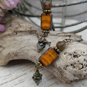 May include: A pair of handcrafted earrings featuring rectangular orange and brown glass beads with a marbled effect. The earrings also include smaller, round brown beads and decorative metal accents. The earrings hang from bronze-colored ear wires.