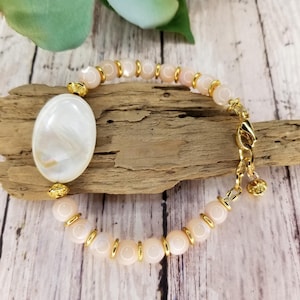 May include: A delicate bracelet with a large oval shell pendant. The bracelet is made of small, pale pink beads with gold accents.